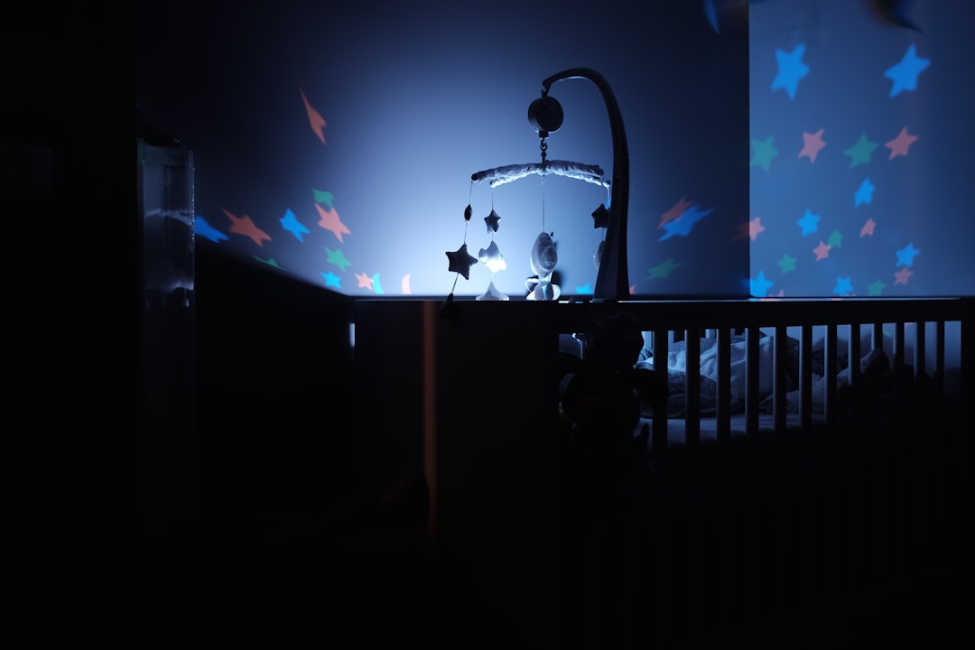 A dimly lit baby crib with a star projection mobile casting colorful star patterns on the walls. The room has a calming, dark blue ambiance, creating a tranquil atmosphere.