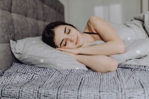 A woman is peacefully sleeping in a bed with gray and white polka dot sheets and a matching pillow. She is lying on her side, resting her head on her hands, with a slight smile on her face. The bed has a tufted headboard.