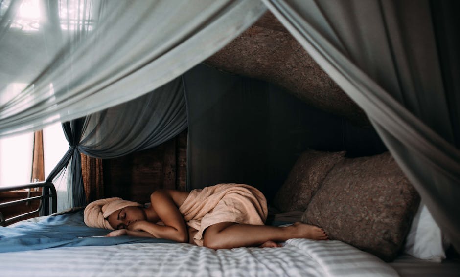 A person wearing a towel turban is lying on their side on a bed under a canopy of sheer curtains, appearing relaxed and peaceful. The room has a cozy, serene atmosphere with soft, muted colors.