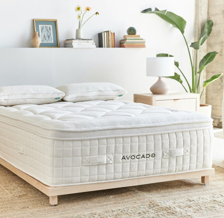 A neatly made bed with a white mattress and two pillows, marked "AVOCADO," stands against a white wall in a bright bedroom. Nearby are a nightstand holding a table lamp and books, a framed photo on the wall, a potted plant, and a natural fiber rug on the floor.