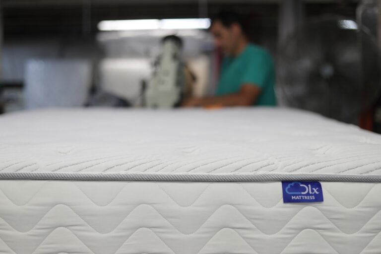 Close-up of a white mattress with grey edges and a blue tag that reads "oLx MATTRESS." A person in a green shirt is blurred in the background, focusing on a machine, suggesting a manufacturing or workshop setting.