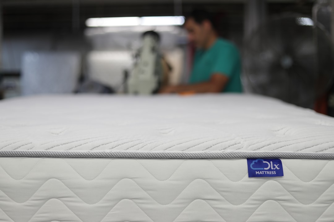 Close-up of a white mattress with grey edges and a blue tag that reads "oLx MATTRESS." A person in a green shirt is blurred in the background, focusing on a machine, suggesting a manufacturing or workshop setting.