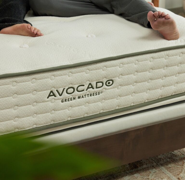 A close-up of an Avocado Green Mattress on a wooden bed frame. Part of a person's legs and feet are visible resting on the mattress. The mattress has a textured fabric surface and a stitched label with the brand name "AVOCADO Green Mattress" in green lettering.