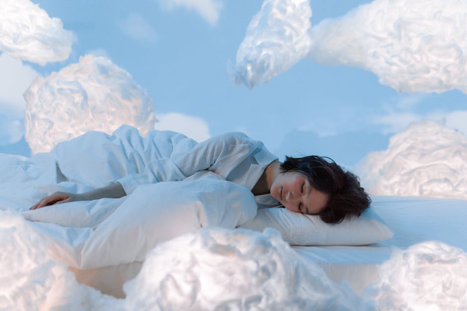 A person sleeps peacefully on a white bed with their head resting on a pillow. The scene is surreal, with large fluffy white clouds surrounding the bed, creating a dreamlike atmosphere. The background is a blue sky with more clouds drifting by.