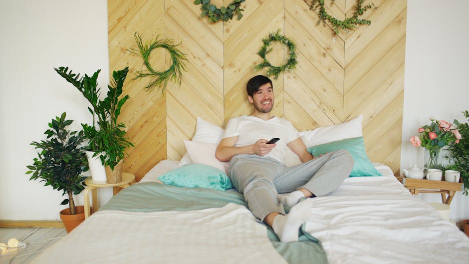 A man lounges on a bed with a chevron-patterned wood headboard, surrounded by green plants and floral decor. He is holding a remote control, wearing a white t-shirt and grey pants, and smiling while looking off to the side.