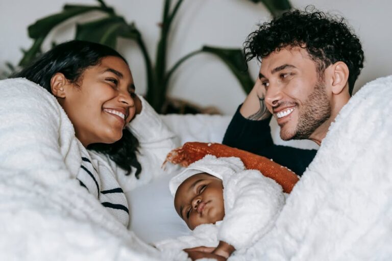 A couple smiles at each other while lying in bed, with a baby sleeping between them. All three are covered in white blankets and appear cozy and happy. There are green plants in the background.