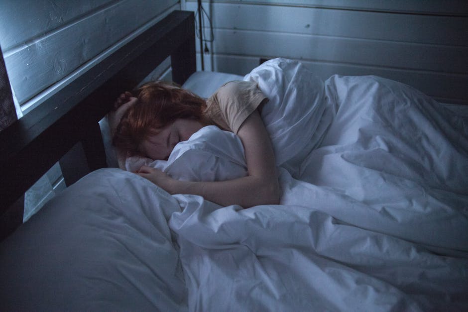 A person with red hair is lying in bed, covered by a white blanket, and resting on white pillows. The room is dimly lit with bluish tones, and a wooden wall can be seen in the background. The person appears to be sleeping or resting.