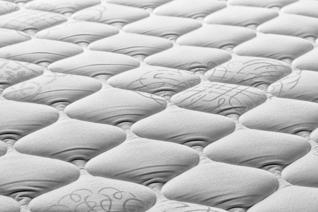 Close-up view of a quilted mattress surface with a tufted pattern. The fabric is light in color and features subtle decorative stitching, creating a series of rounded, cushion-like shapes across the mattress.