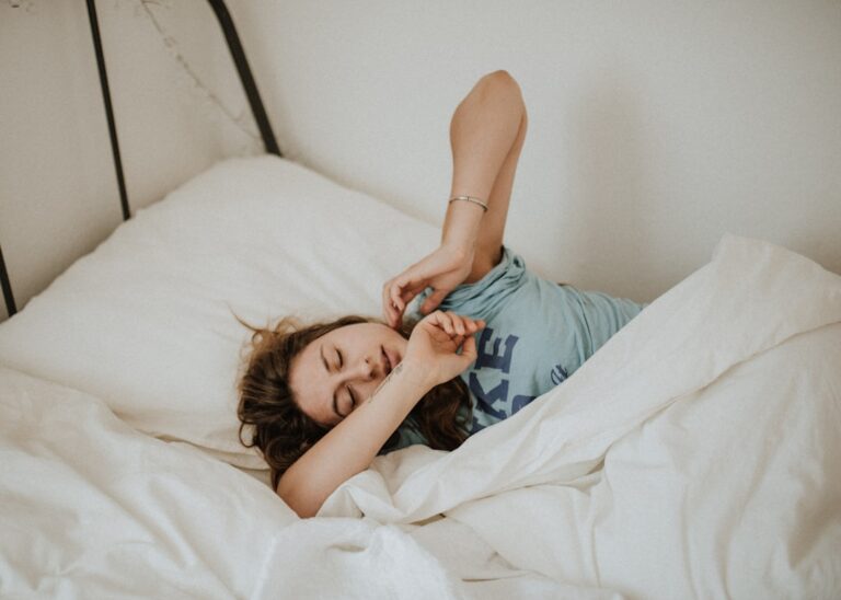 A person with long hair is lying in bed, stretching with their eyes closed. They are on white bedding and wearing a light blue t-shirt. The scene suggests a relaxed morning or a moment of waking up.