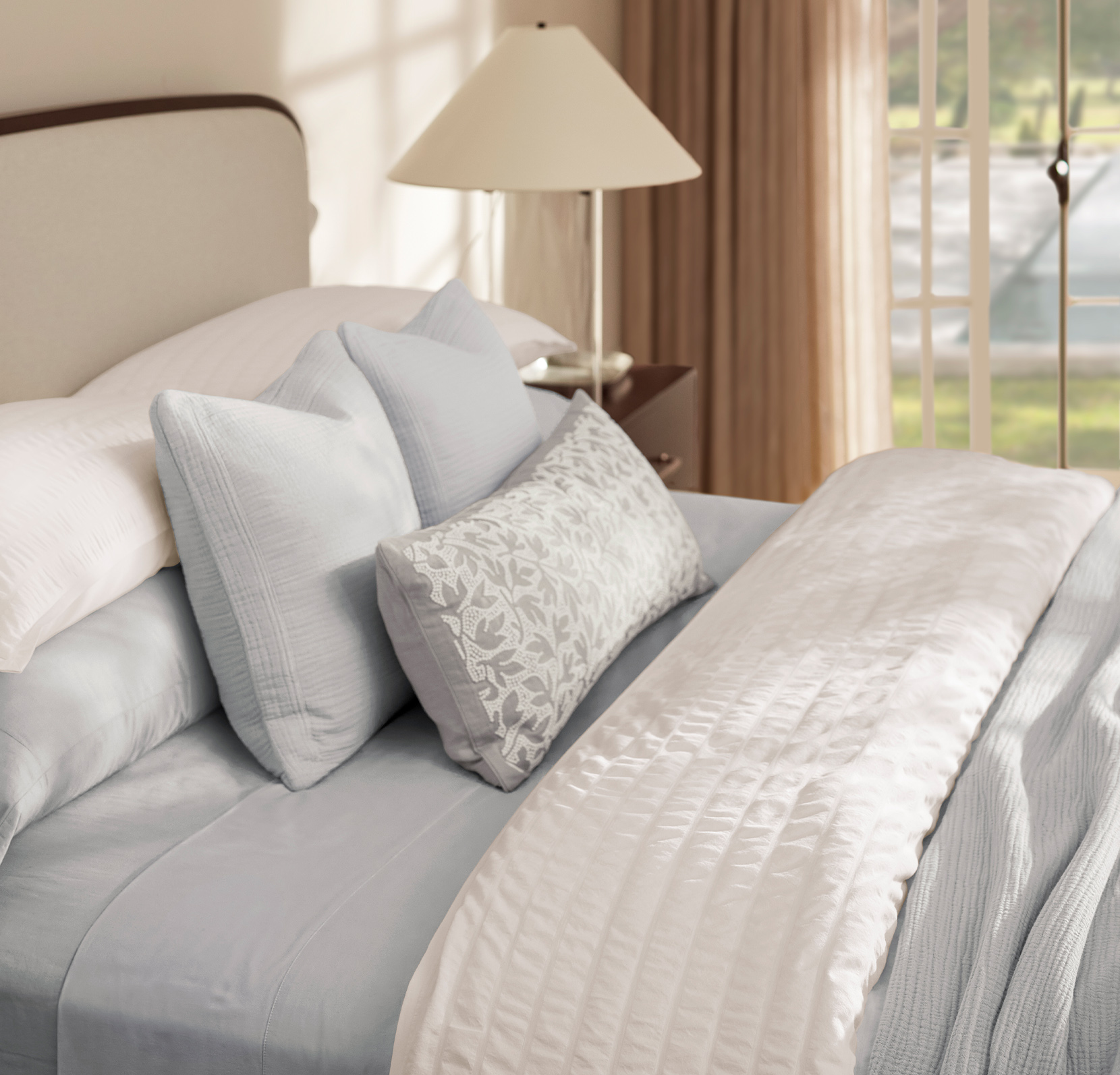 A neatly made bed adorned with light blue and white bedding. Several pillows are arranged at the head of the bed, including two large blue ones and a smaller one with a floral pattern. A bedside table with a lamp is situated to the right, near a window with open curtains.