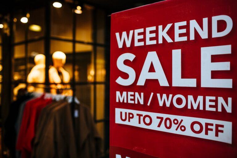 A store window displays mannequins dressed in clothing. In the foreground, a red sign promotes a "Weekend Sale" with discounts of up to 70% off for men's and women's apparel.