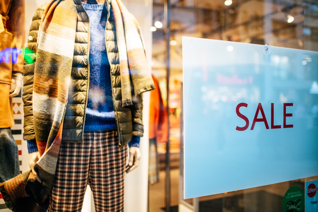 Mannequin wearing winter clothing, including a puffer jacket, blue sweater, scarf, and plaid pants, displayed in a store window. A large white sign with red letters that read "SALE" is posted on the glass, indicating a promotion or discount event inside the store.