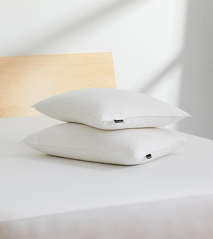 Two white pillows stacked on top of each other on a neatly made bed with white sheets. The headboard is wooden, and soft natural light illuminates the scene from the right.