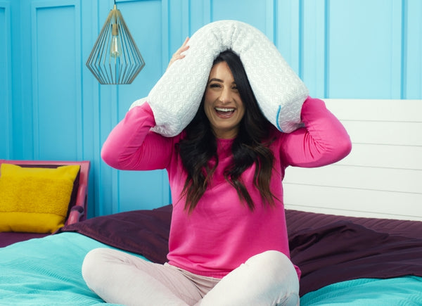 A woman with long dark hair and a bright smile sits cross-legged on a bed with teal and purple bedding, holding a pillow over her head. She is wearing a pink long-sleeve shirt. The background features blue paneled walls, a hanging light fixture, and colorful pillows.