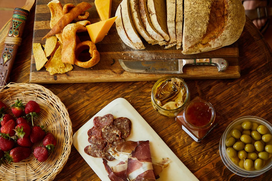 A rustic table setting featuring sliced bread on a wooden cutting board with a knife, assorted cheeses, dried fruits, a basket of strawberries, cured meats on a white board, a jar of artichoke hearts, a jar of sauce, and a bowl of green olives.