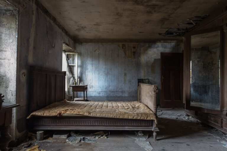 A dilapidated, dimly-lit room with peeling paint and damaged walls. A dusty, worn-out bed with a headboard is in the center, flanked by a small bedside table. Debris and broken furniture are scattered on the floor, and a large mirror stands against the wall.