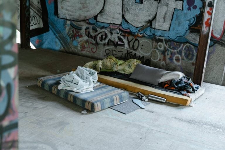 A makeshift sleeping area under a graffiti-covered bridge includes a mattress and a sleeping mat with blankets and pillows. Surrounding the bedding are scattered personal items and debris. The setting appears to be an urban environment.