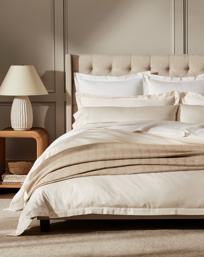 A neatly made bed with soft beige and white bedding, including multiple pillows and a textured blanket, stands in a well-lit bedroom. A tufted headboard is present, and a wooden nightstand with a lamp and decorative items is next to the bed.