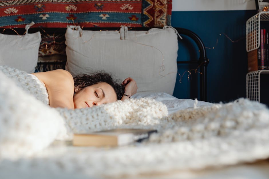 A person with curly hair sleeps peacefully in a cozy bed with a knitted blanket. There are pillows behind them and a decorative rug hangs on the wall above. A book and glasses lie on the bed near the person. Shelves with books are visible in the background.