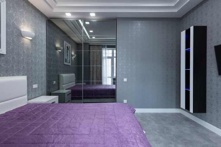 A modern bedroom with a purple quilted bed, grey walls, and a mirrored wardrobe. There is a white headboard, a mounted shelf unit with blue lights, and a wall-mounted TV. Ceiling lights illuminate the room, reflecting off the mirrors, giving a spacious feel.