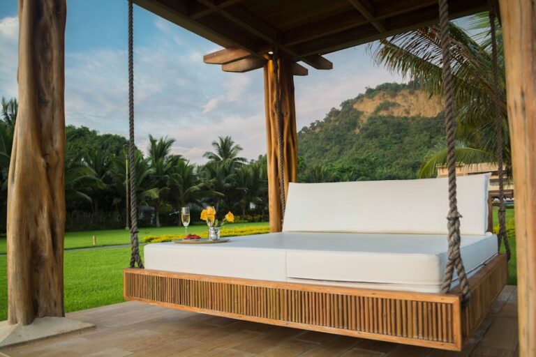 A luxurious outdoor daybed with white cushions is suspended by ropes under a wooden pergola, set in a lush green garden with palm trees and hills in the background. Two glasses of white wine and a small platter of snacks sit on a side table nearby.