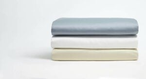 A neatly folded stack of three bed sheets is displayed against a plain white background. The sheets are arranged in a tiered manner, with the top sheet in a light blue color, the middle sheet in white, and the bottom sheet in a cream color.