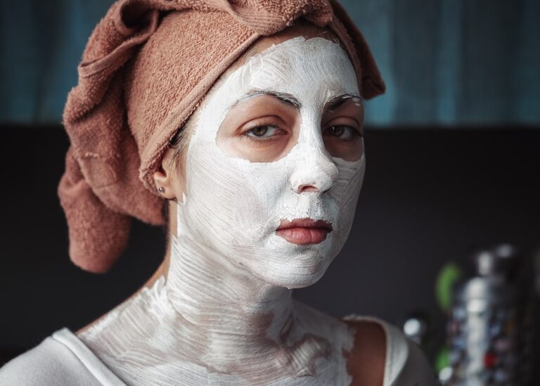 A person with a beige towel wrapped around their head has a white facial mask applied to their face and neck, looking directly at the camera with a calm expression. The background is blurred, suggesting an indoor setting.