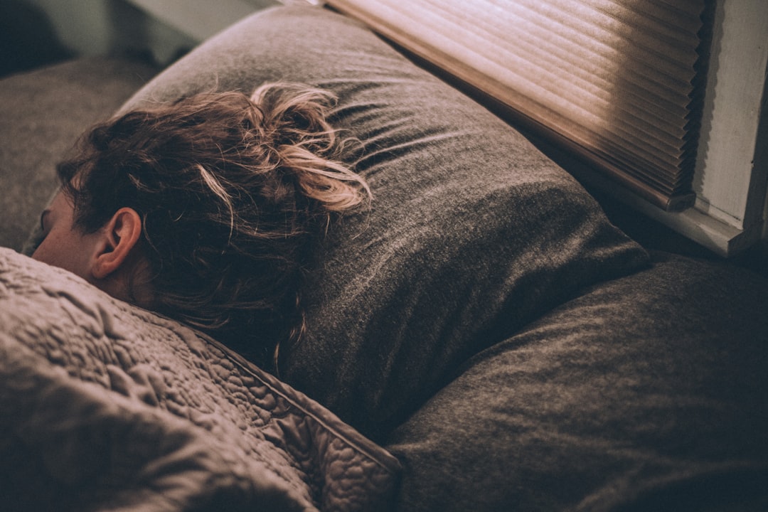 A person with light brown hair is sleeping on a bed, facing away from the camera. They are covered with a gray blanket, with a gray pillow under their head. A window with a closed blind is partially visible in the background.