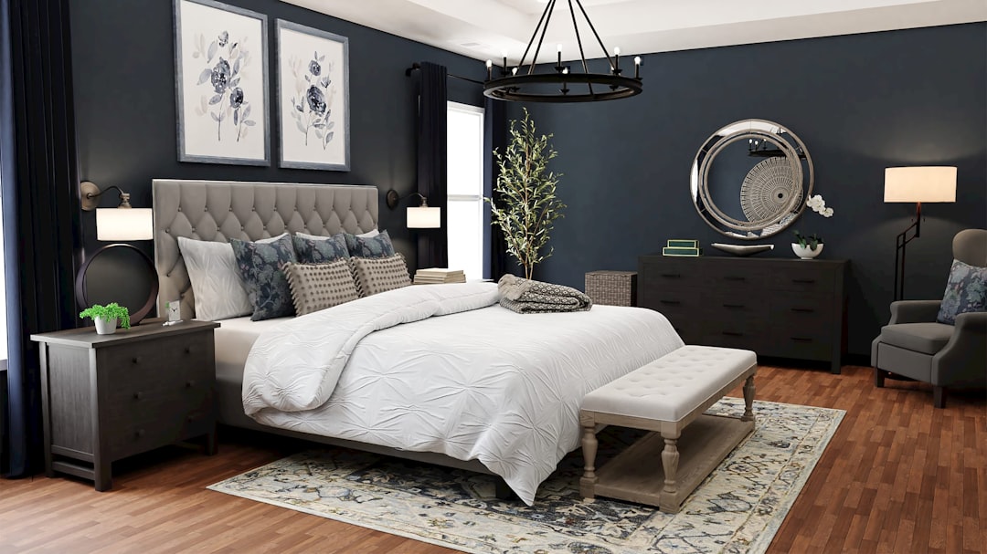 A modern bedroom with dark blue walls, wooden floors, and a large bed with a tufted headboard. The bed has white bedding with blue and gray pillows. There are matching nightstands, a round mirror above a dresser, wall art, a chandelier, and a upholstered bench.