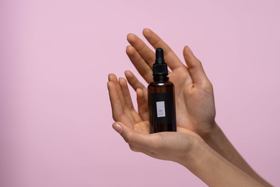 Two hands gently hold a small brown glass bottle with a dropper against a pink background. The bottle has a white label with the text "Oil Dropper" visible. The composition highlights the product elegantly.