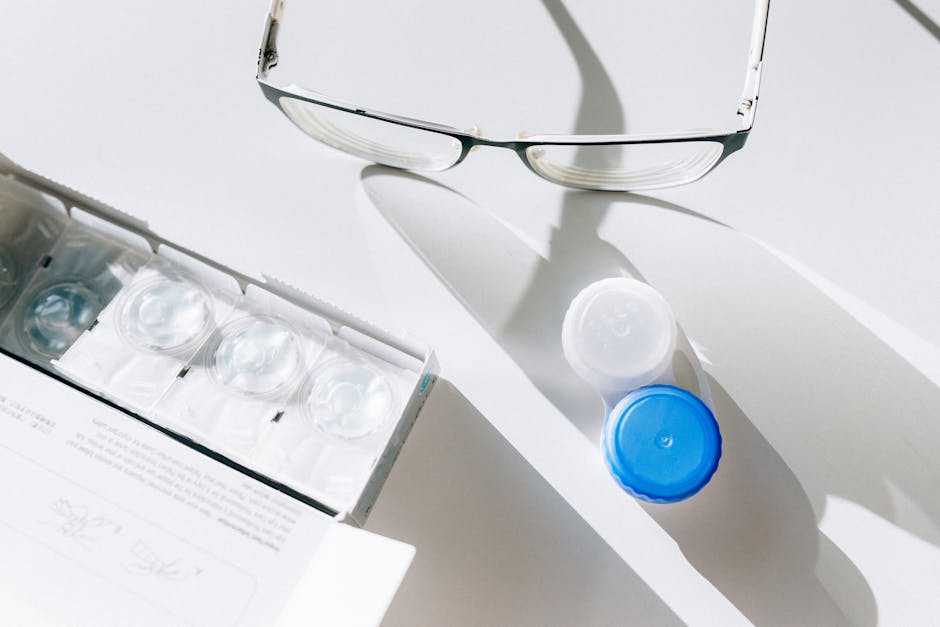 A pair of eyeglasses, an open contact lens case with one blue cap and one white cap, and several blister packs containing contact lenses are laid out on a white surface with shadows.