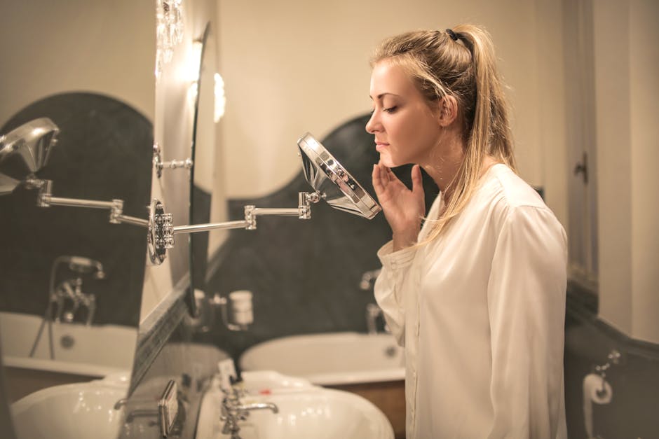 A woman with blond hair in a ponytail stands in front of a bathroom mirror. She is wearing a light, white blouse and looking at her reflection in a small circular magnifying mirror, touching her face gently. The bathroom has two sinks in the background.