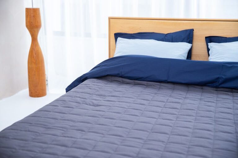 A neatly made bed with a wooden headboard features two pillows with blue pillowcases and a navy blue and gray bedspread. A tall, wooden lamp stands beside the bed against a background of white curtains.