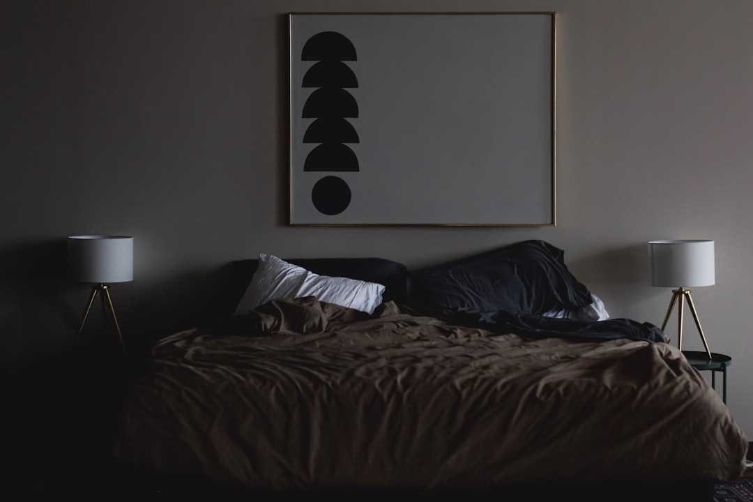 A dimly lit bedroom with an unmade bed featuring dark sheets and pillows. On the wall above the bed hangs a framed abstract art piece with black geometric shapes. Two bedside tables, each with a white lampshade, sit on either side of the bed.