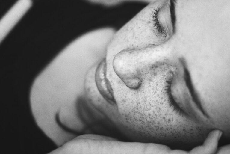 Close-up black and white photo of a person with freckles lying down with eyes closed, their face in peaceful repose. The image focuses on the serene expression and detail of the person's facial features.