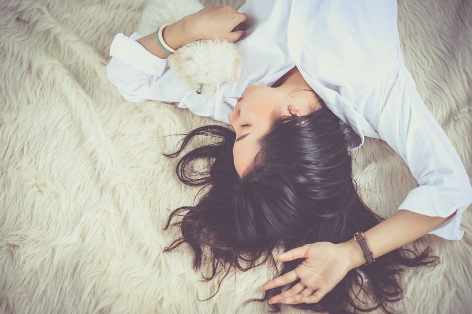 A person with long dark hair lies on a fluffy cream-colored rug, wearing a white shirt. They are holding a small white dog close to their face while resting on their back, appearing relaxed and peaceful. The dog looks cozy and nestled against the person's chest.