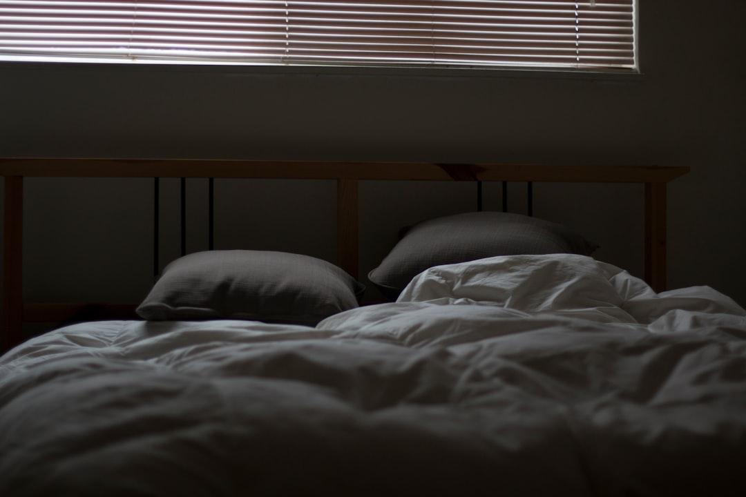 A dimly lit bedroom showing a bed with two pillows and a rumpled white comforter. A window with closed blinds lets in a small amount of light, casting shadows in the room. The headboard of the bed is simple and wooden.