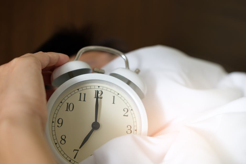 A hand reaching out to turn off a white analog alarm clock. The clock face shows the time is 7:00. The person is lying in bed, partially covered by a white blanket, suggesting they are waking up. The background is blurred.