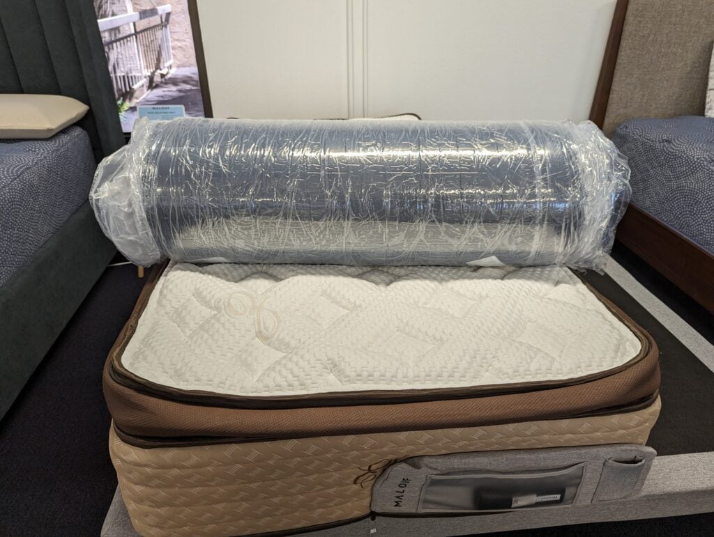 A rolled-up, vacuum-sealed mattress lies atop a regular mattress on display in a showroom. The rolled mattress is wrapped in clear plastic, while the regular mattress has a white quilted top and a brown fabric border with a visible side handle.