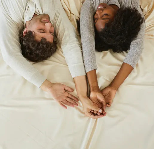 A couple lies on a cream-colored bed, facing each other with arms outstretched and hands intertwined. They are smiling warmly at each other, wearing light-colored clothing, and the scene exudes a sense of joy and intimacy.