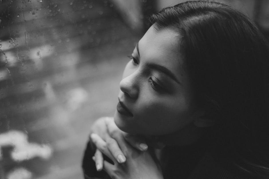 Black and white photo of a woman with long hair, leaning her chin on her hands and gazing out a rain-streaked window. Her expression is introspective, contributing to the image's contemplative mood.