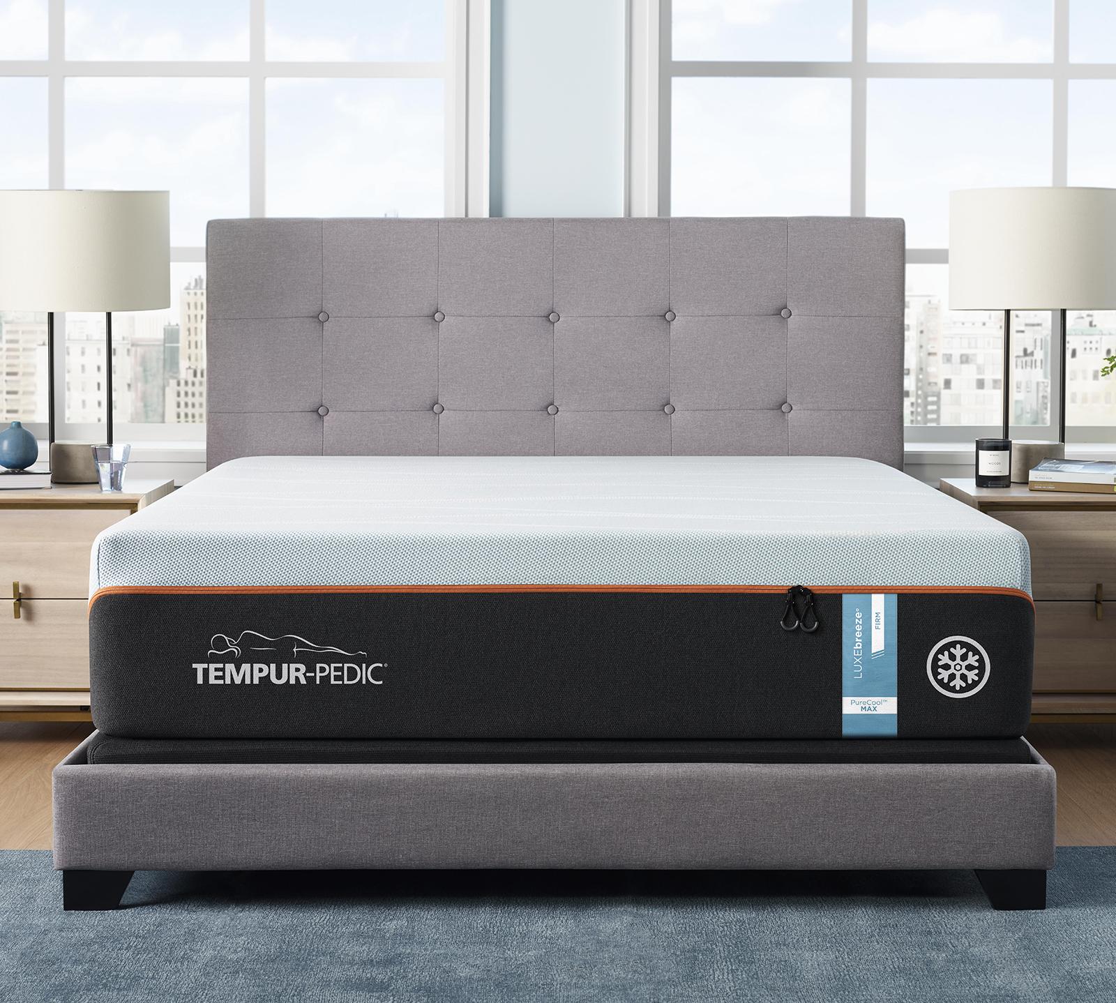 A neatly made bed with a grey upholstered headboard and frame features a Tempur-Pedic mattress labeled with cooling technology. The bed, flanked by two wooden nightstands with lamps and decor, sits beneath a large window displaying a cityscape view. Wondering how much is a Tempurpedic bed?