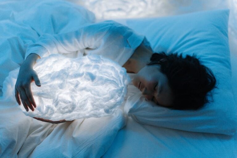 A person lies in bed, asleep, embracing a glowing cloud-shaped object. The scene is bathed in soft blue light, creating a surreal, dreamy ambiance. The person is dressed in white, which adds to the ethereal quality of the image.