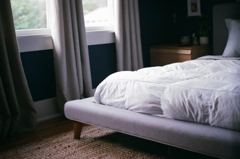 A cozy bedroom featuring a modern, low-profile bed with a white comforter. The room has dark walls, large windows with curtains, a light wooden side table, and a woven rug on a wooden floor, creating a serene and inviting atmosphere.