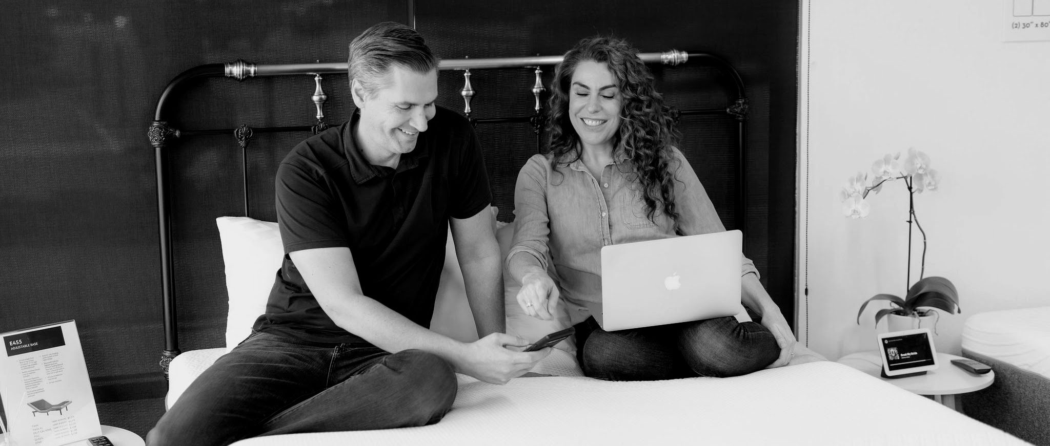 A black and white photo of two people sitting on a bed. The person on the left holds a phone while the person on the right, who has a laptop on their lap, points at the phone screen. They are both smiling.