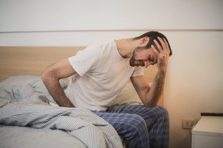 A man wearing a white t-shirt and blue plaid pajama pants sits on the edge of a bed, holding his head with one hand and his stomach with the other, appearing to be in pain. The room has a neutral-colored wall and a nightstand beside the bed.