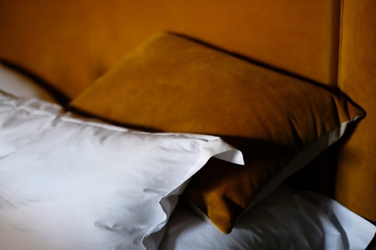 A cozy bedroom scene featuring a mustard yellow pillow propped against a yellow upholstered headboard. A crisp white pillow and part of the white bed sheets are also visible, contrasting with the warm tones of the yellow elements.