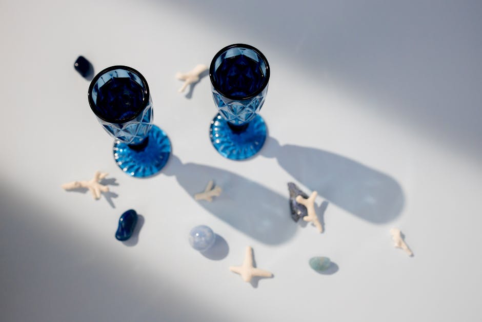 Top view of two blue crystal glasses on a white surface, surrounded by small seashells and rocks. The glasses cast long shadows and the geometric pattern on them creates intricate light reflections.