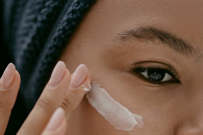 A close-up shot of a person gently applying a smear of white cream to their cheek. The person has neatly shaped eyebrows and a calm expression. Their well-manicured nails and the texture of the cream are visible in detail.