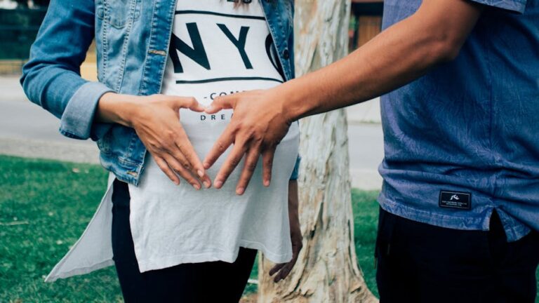 A close-up of a couple standing outdoors, with their hands forming a heart shape over the woman's pregnant belly. The woman is wearing a white shirt, partially covered by a denim jacket, and the man is dressed in a blue shirt.